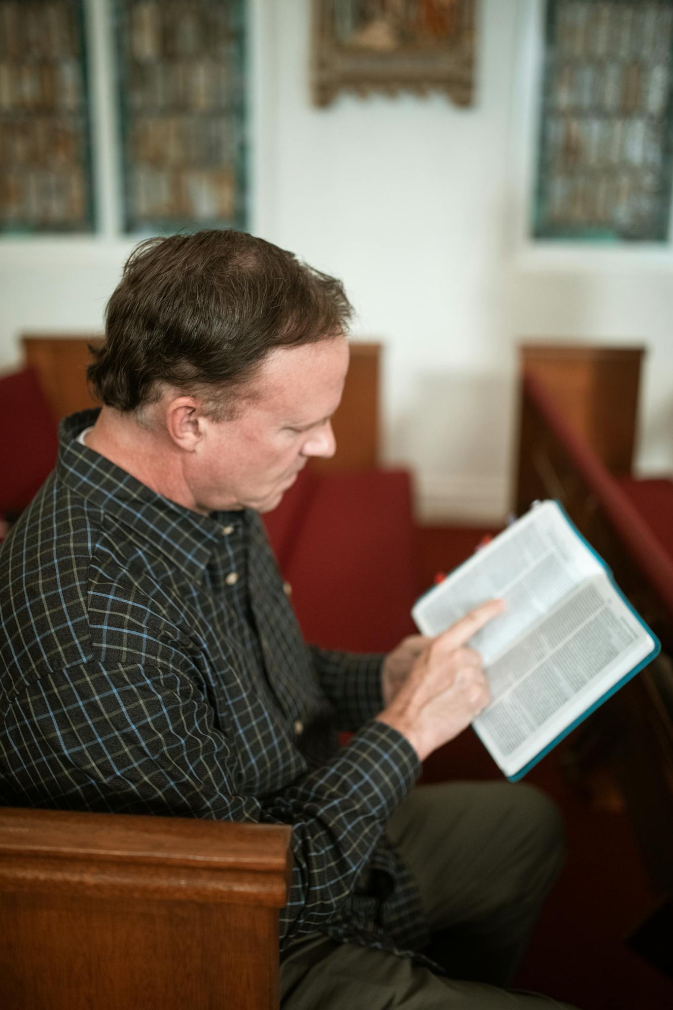 Side view of concentrated senior male in casual clothes reading holy book while sitting on bench in church