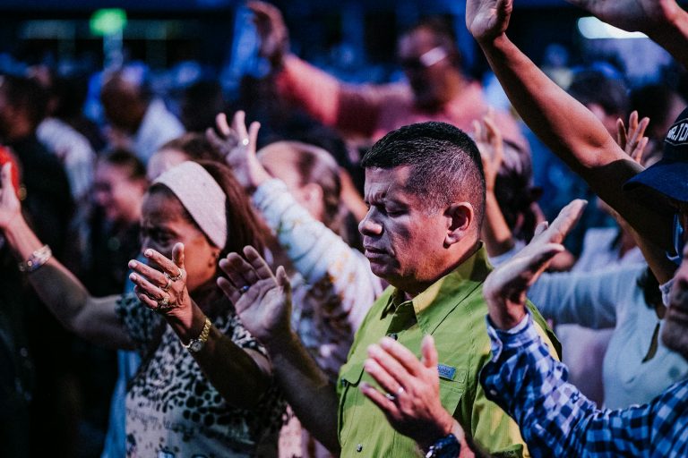 A vibrant crowd of diverse adults worshipping with raised hands indoors.